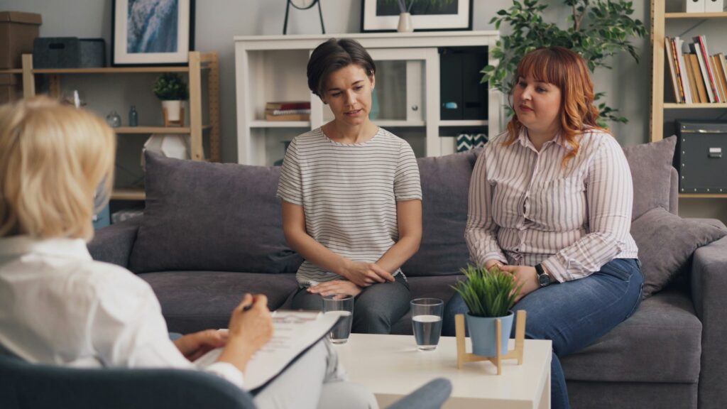 A couple sitting on a couch and talking to a woman with a clip board